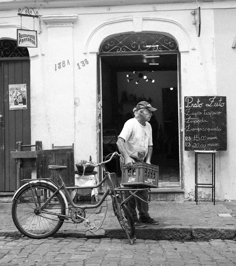 Elderly man stands beside bicycle on cobblestone street in São João del Rei.